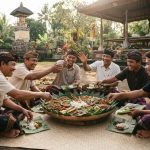 Sejumlah pria Bali tersenyum dan makan bersama dari piring besar daun pisang (megibung) di halaman pura tradisional