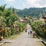 Pemandangan jalan utama Desa Penglipuran, Bali, yang bersih dan asri, diapit oleh gerbang tradisional Bali, pepohonan tropis, dan rumah-rumah beratap ijuk dengan latar belakang perbukitan hijau