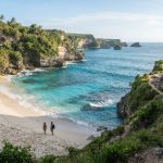 Sepasang pejalan kaki berjalan di sepanjang pantai terpencil di Bali, dengan tebing hijau yang dramatis dan air biru kehijauan yang jernih di kejauhan