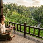 Seseorang sedang bermeditasi santai di balkon kayu dengan pemandangan lanskap terasering sawah hijau dan hutan tropis yang asri di Ubud, Bali.