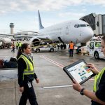 Petugas ground handling bandara sedang menggunakan perangkat tablet cerdas di area apron dengan latar belakang pesawat terbang dan peralatan operasional bandara.